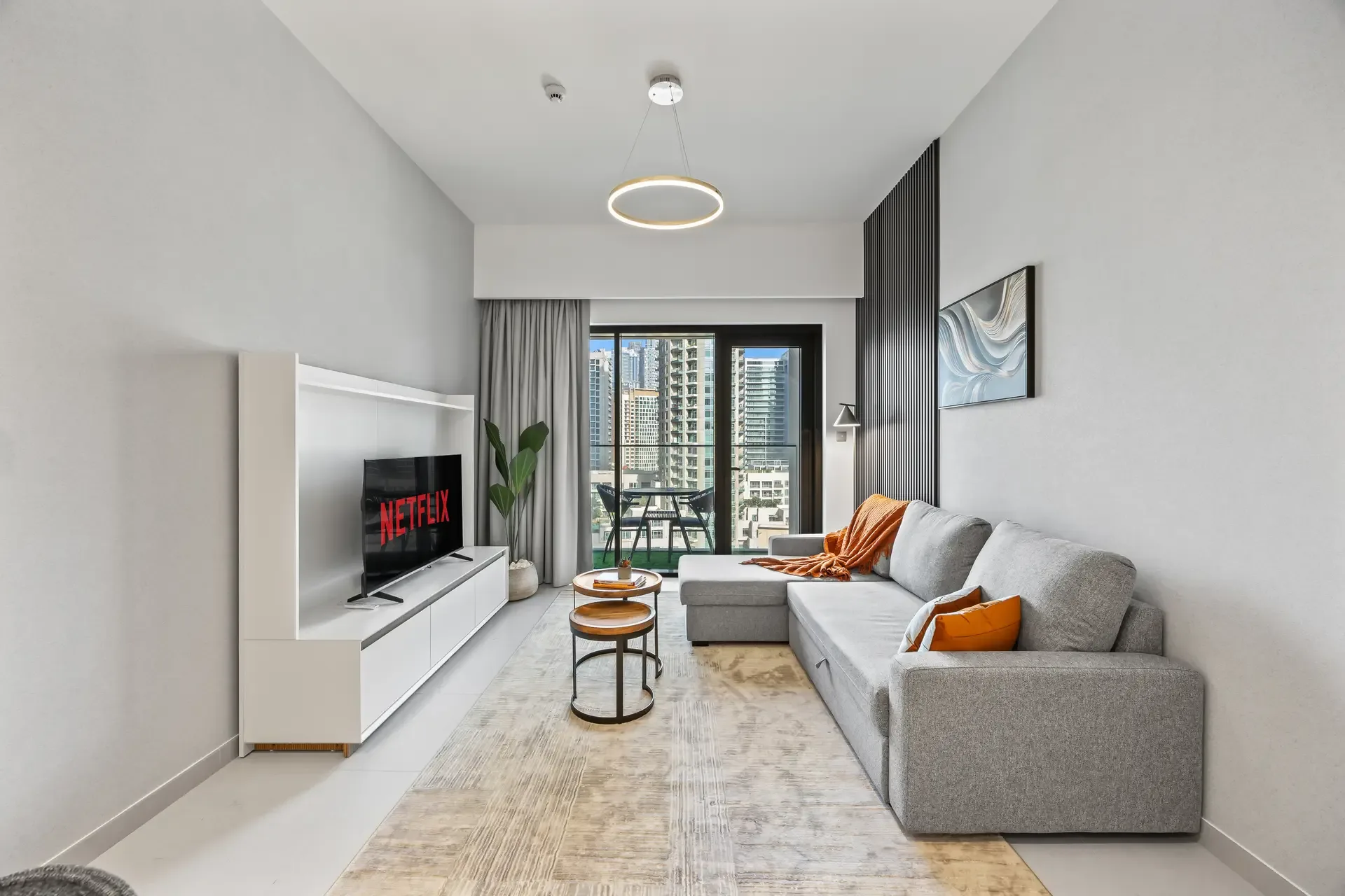 Modern living room with a gray sectional sofa, orange cushions, and a white media console hosting a Netflix TV. A balcony with city views is visible outside.