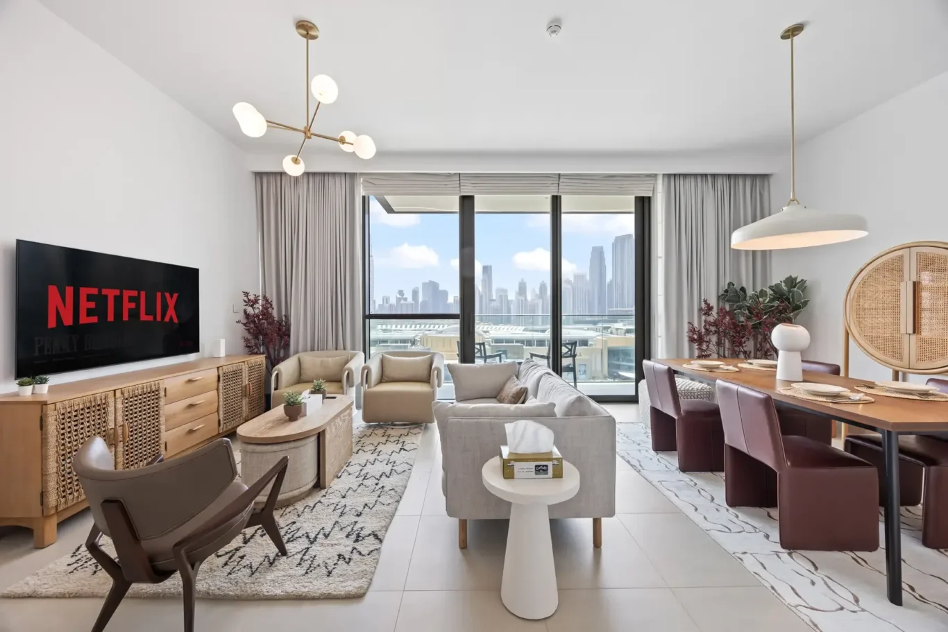 Bright open-plan living room with a Netflix TV, beige sofa, wicker cabinet, and large windows overlooking a city skyline.