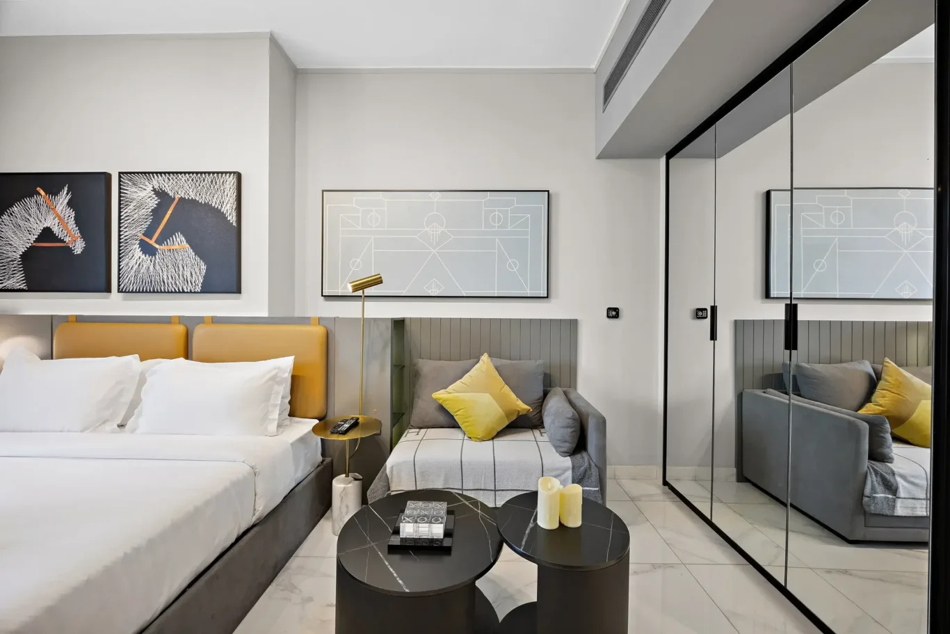 Modern hotel room featuring a white bed with a mustard headboard, gray sofa, abstract line-art paintings, and a mirrored wardrobe and glass wall.