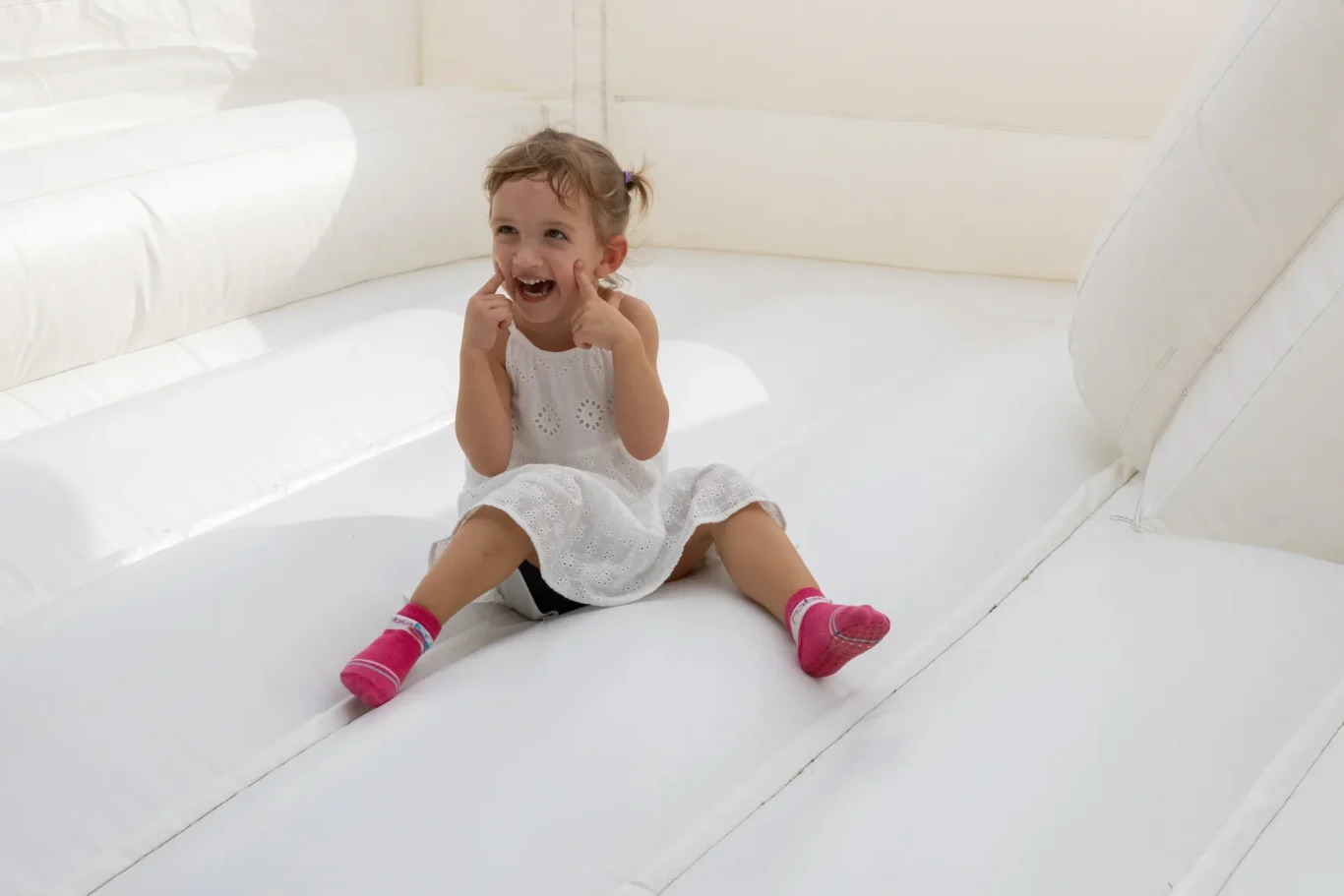 Smiling young girl in a white dress sits on a white inflatable surface, pink socks visible on her feet.