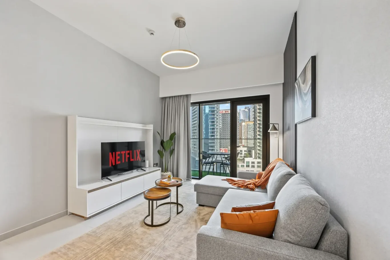Modern living room with a gray sofa, orange cushions, and a TV showing Netflix, plus white media unit and round nesting tables. A glass balcony door reveals a city skyline.