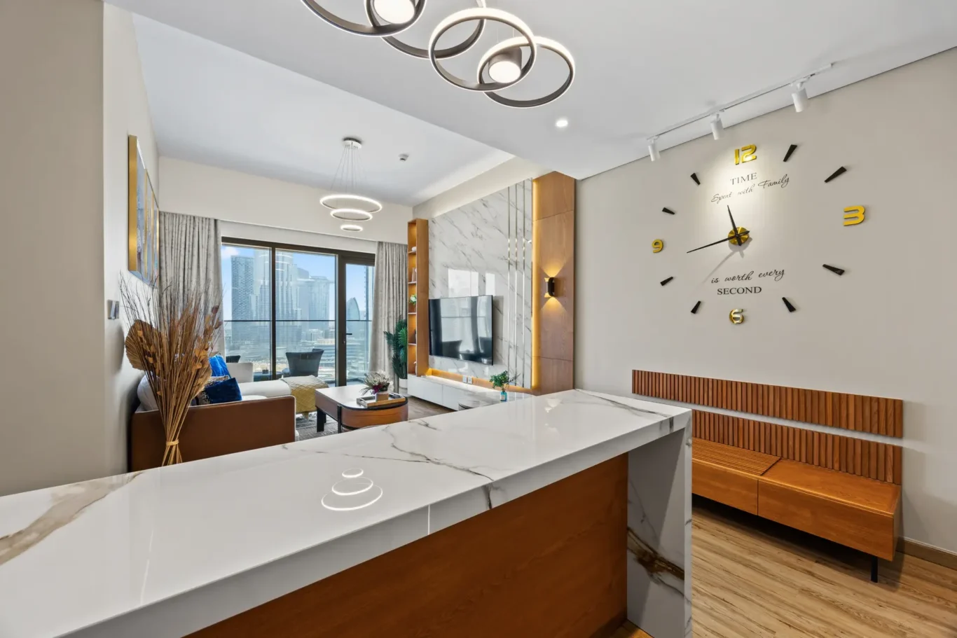 Modern living room with marble countertops, wooden storage bench, and a large decorative wall clock with gold numerals; city skyline visible through glass doors in the background.