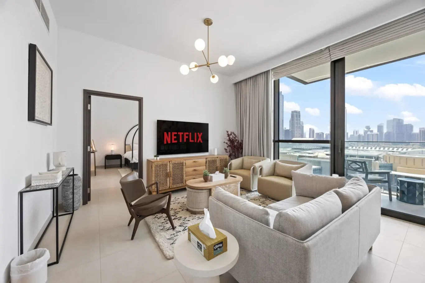 Living room with a beige sofa set, TV showing Netflix, and a wooden sideboard facing a large window balcony with city skyline view.