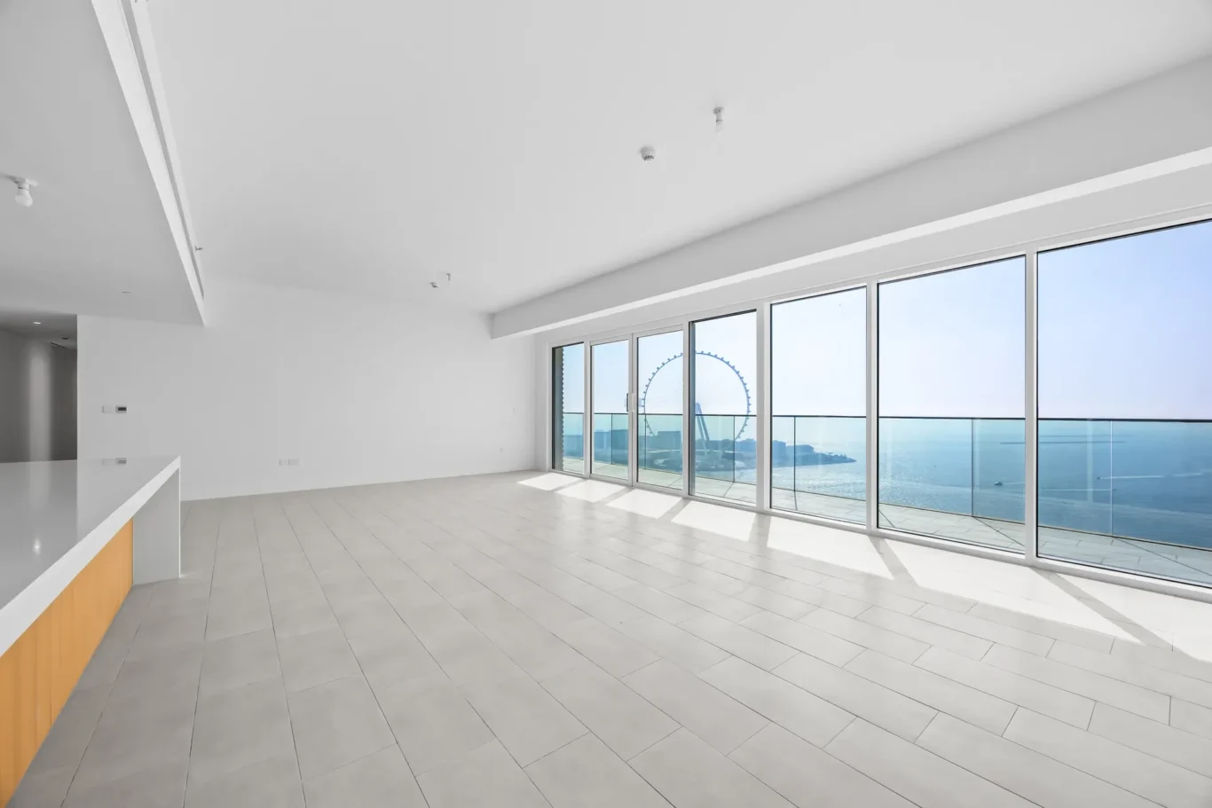 Bright empty living space with floor-to-ceiling glass windows showing the ocean and a distant ferris wheel outside.