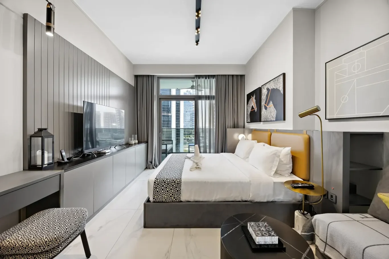 Modern hotel room with a king bed, gray headboard, wall-mounted TV, and long gray storage cabinet facing a balcony view of city skyscrapers.