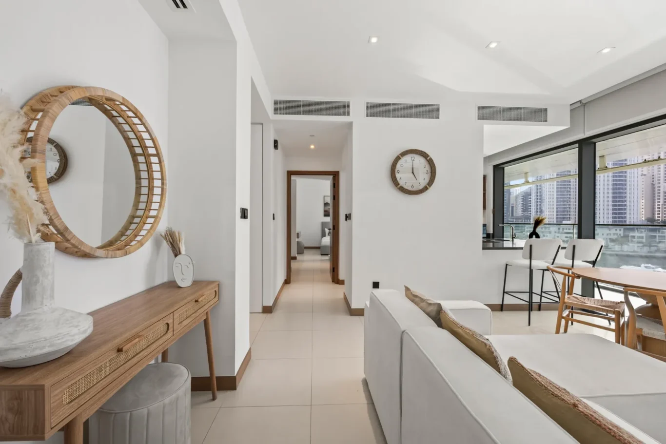 Bright modern living area with white walls, a round wicker mirror above a wood console, and large windows showing the city skyline.