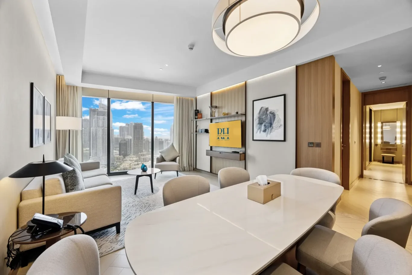 Spacious hotel living area with a large white dining table, beige chairs, a sofa, and floor-to-ceiling windows showing a city skyline.