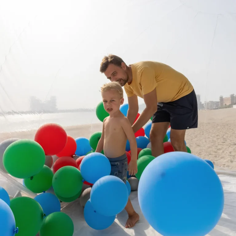A man and a young boy play among colorful balloons (green, blue, red) inside a clear inflatable structure on a sandy beach near the water.