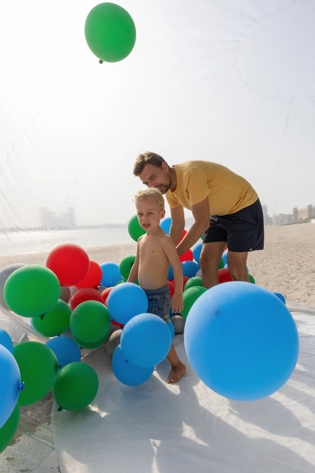 A man and a young boy play among colorful balloons (green, blue, red) inside a clear inflatable structure on a sandy beach near the water.