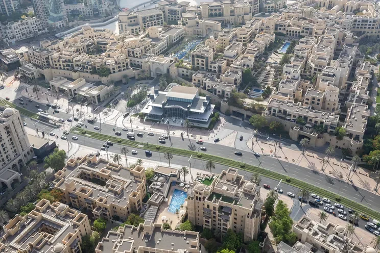 Aerial view of a dense residential complex with beige apartment buildings, a central plaza, and a busy boulevard lined with trees.