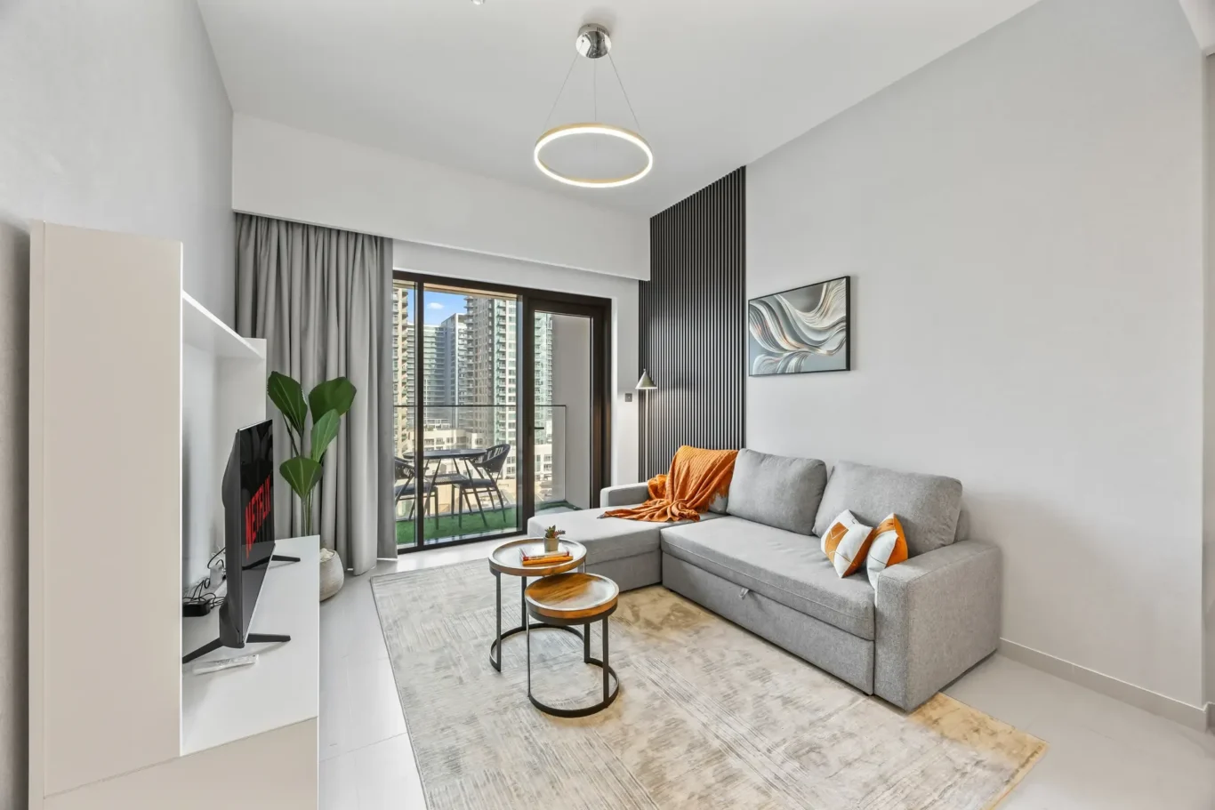 Modern living room with a gray sectional, orange throw pillows, and two round nesting coffee tables by a balcony door overlooking city high-rises.