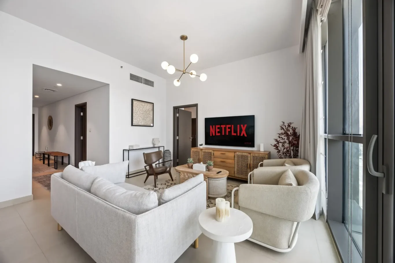 Bright living room with a TV showing the Netflix logo, beige sofas, and a wooden cabinet by a large window to the right.