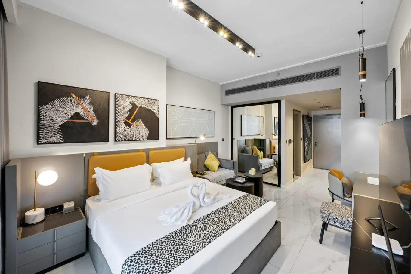 Luxurious hotel room with a king bed, mustard headboard, geometric runner, and a seating area against gray walls with art above the bed.