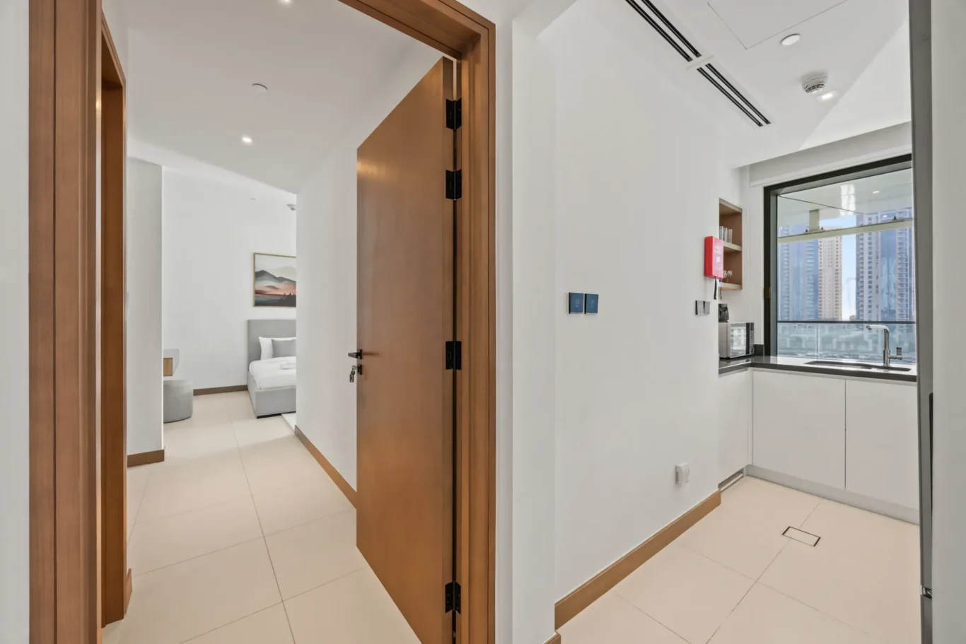 Wide entrance hallway to a modern apartment with open wooden doors revealing a bedroom, and a white kitchen to the right with a city view through a large window.