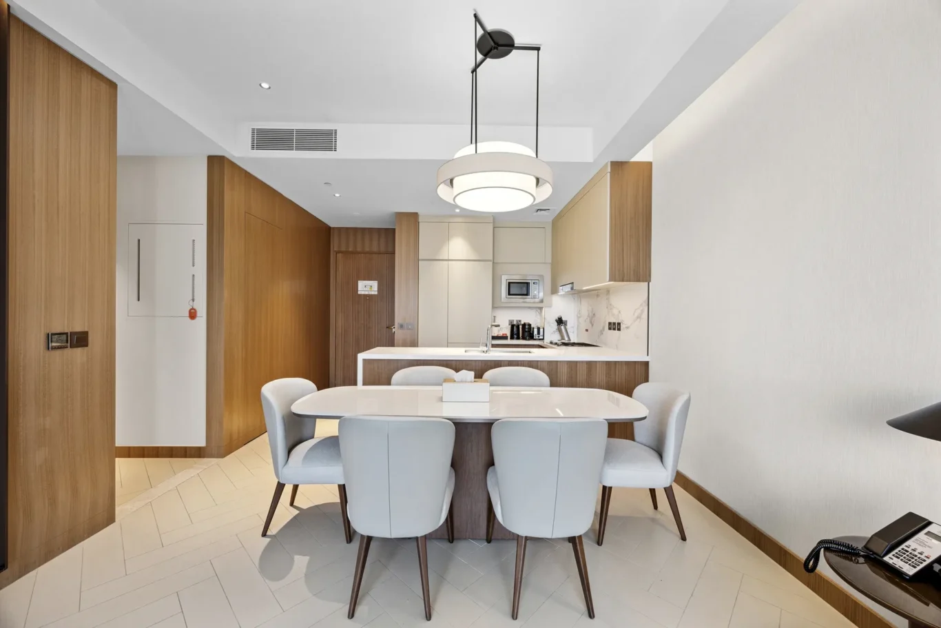 Open-plan kitchen and dining area with a white oval table, six grey upholstered chairs, wood cabinetry, and a large circular pendant light.