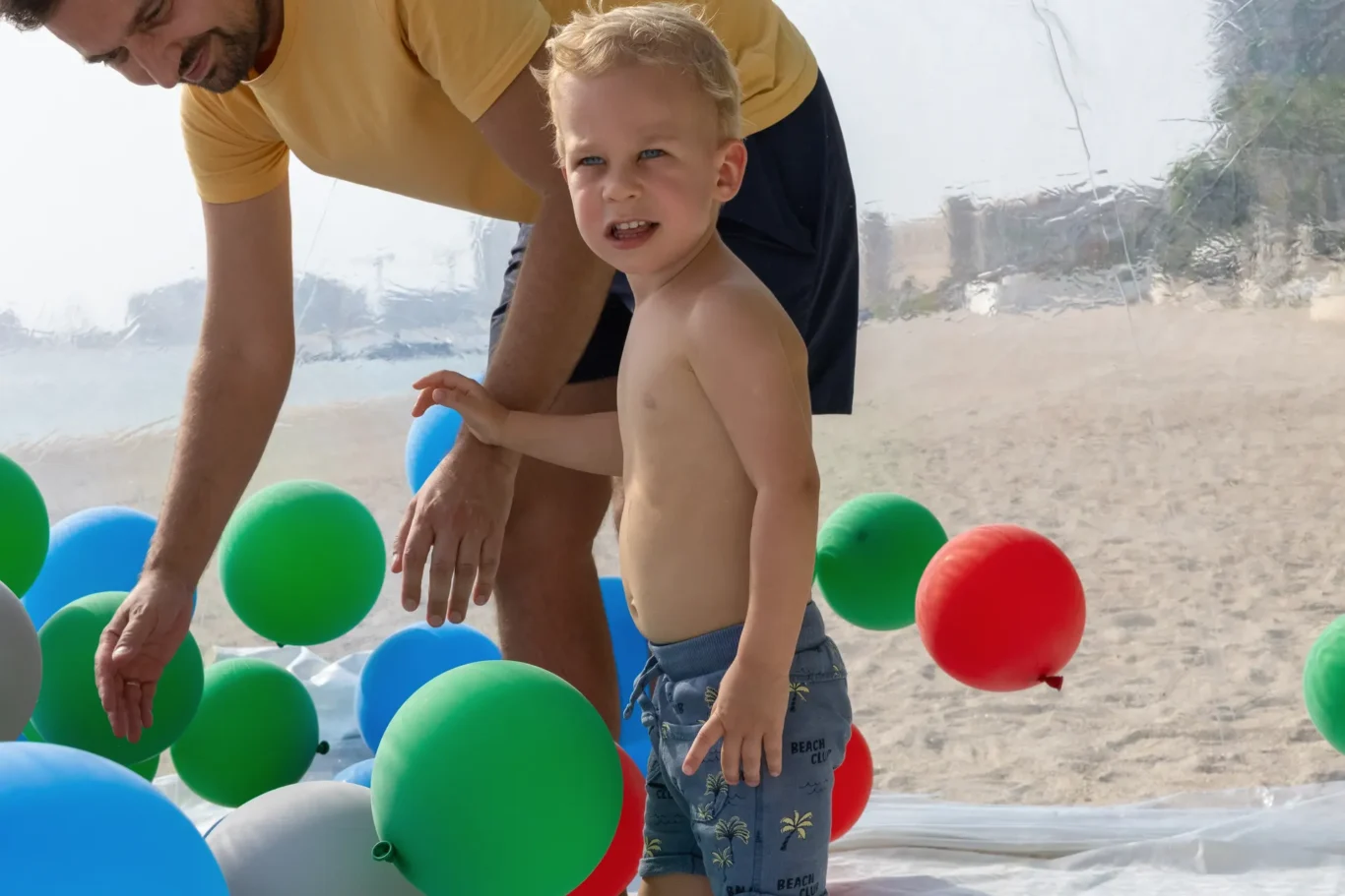 Young shirtless boy at the beach surrounded by colorful balloons, with an adult bending over beside him