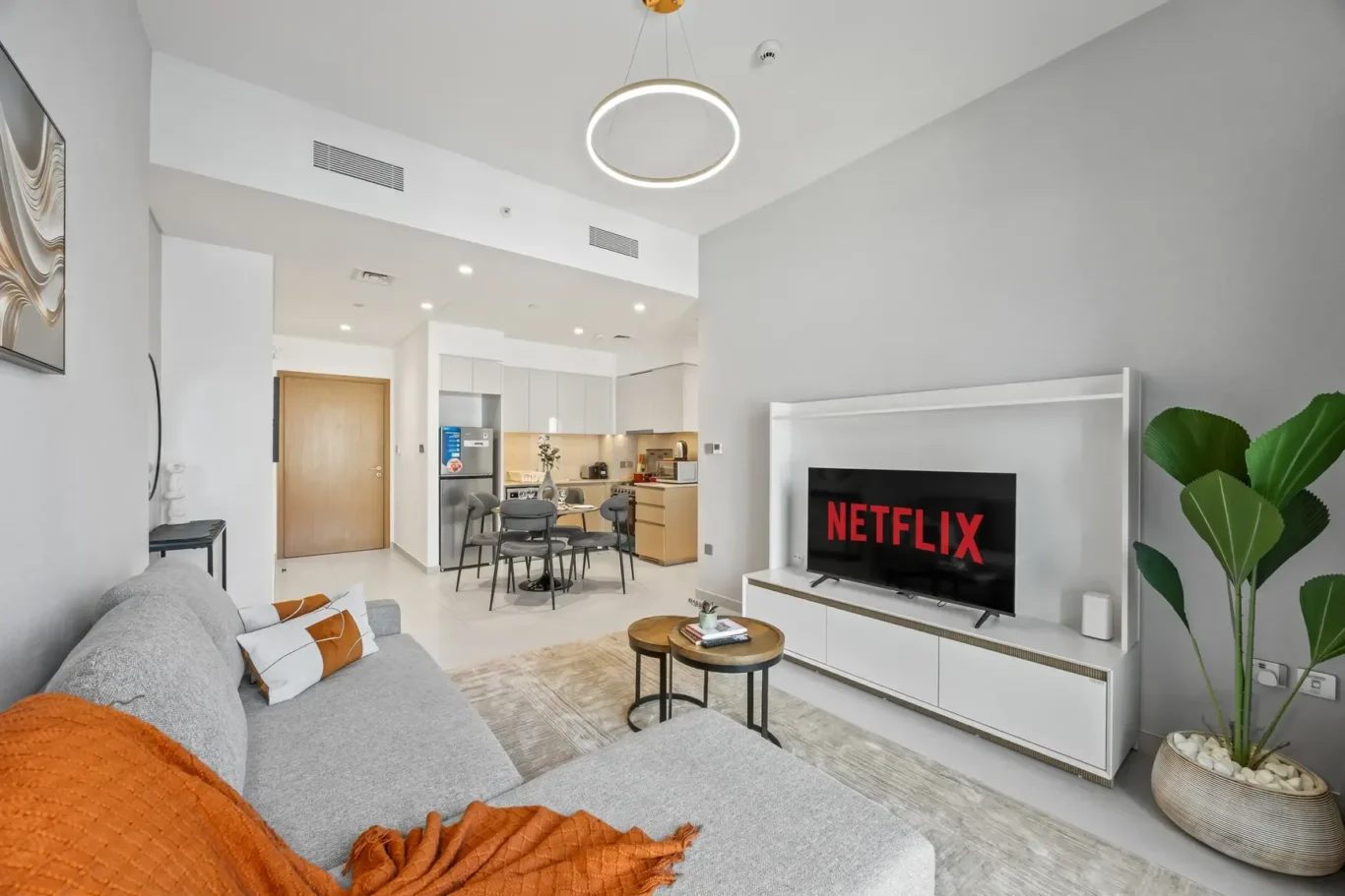 Open-plan living room with a grey sectional sofa, orange throw, round coffee table, and a TV displaying Netflix on a white entertainment unit.