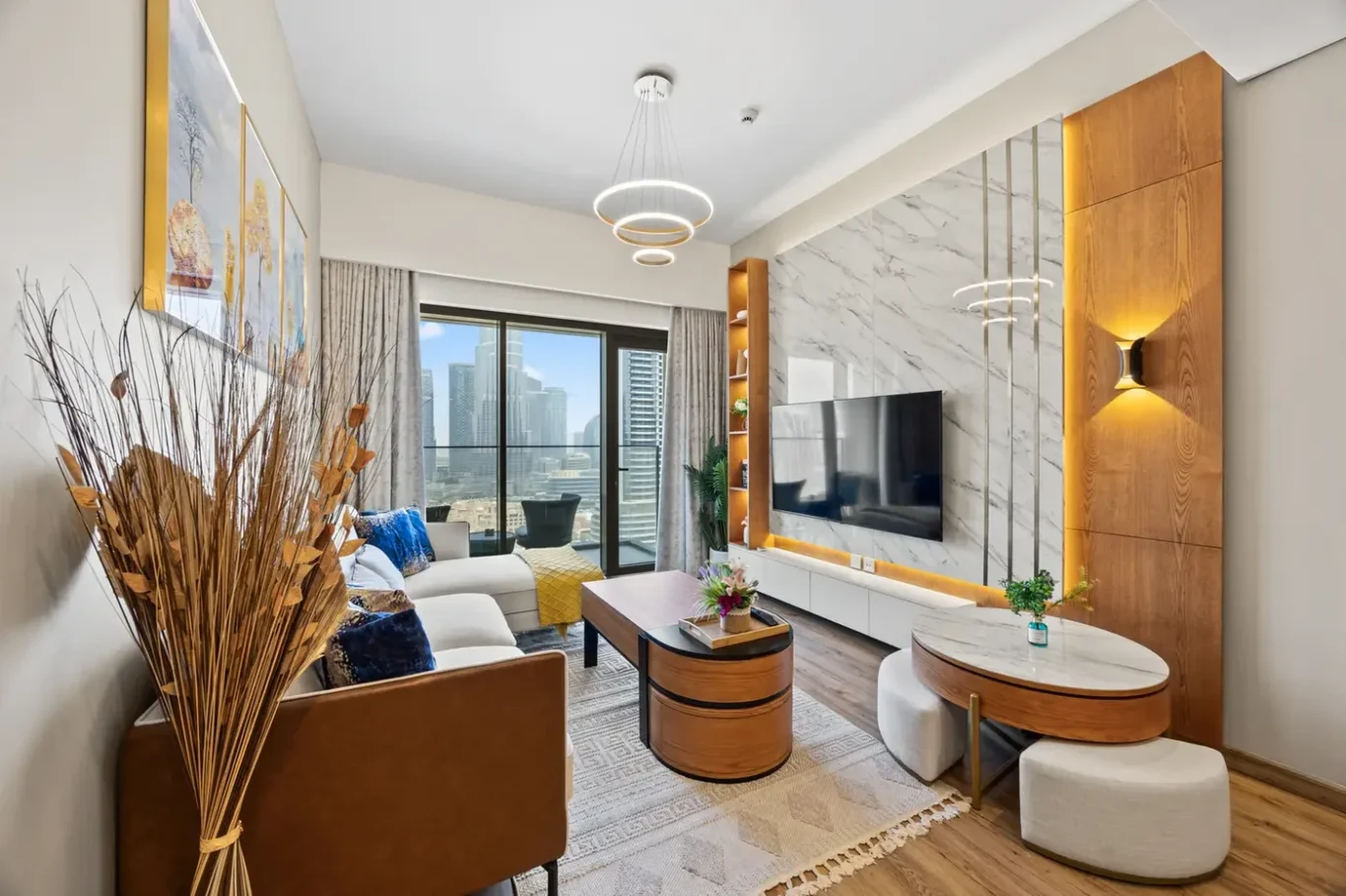 Bright contemporary living room with a white sofa, round wooden coffee tables, and a marble TV wall overlooking a city skyline through large windows.