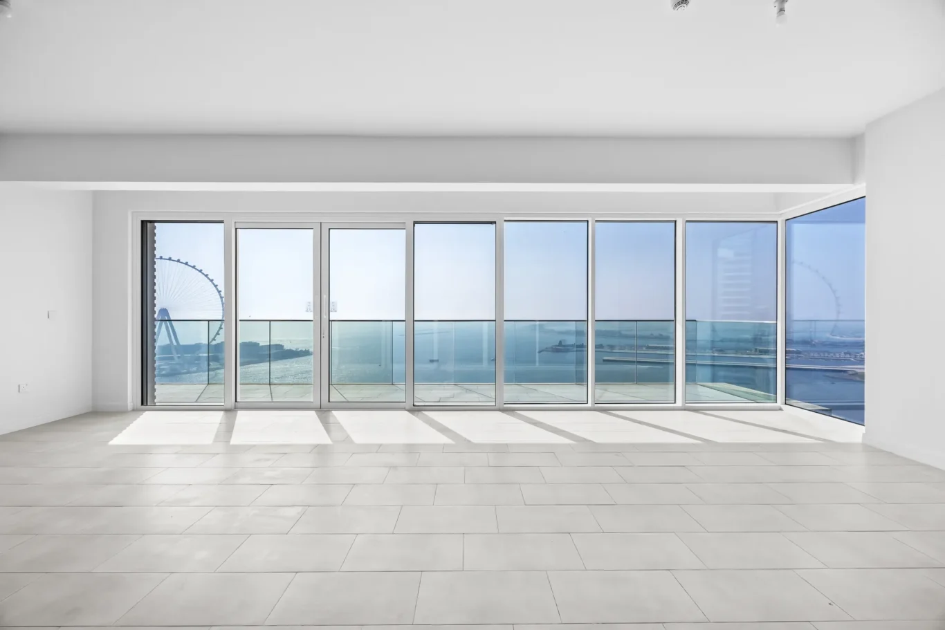 Bright empty room with wide glass doors revealing an ocean view and a partial Ferris wheel outside on the balcony rail.