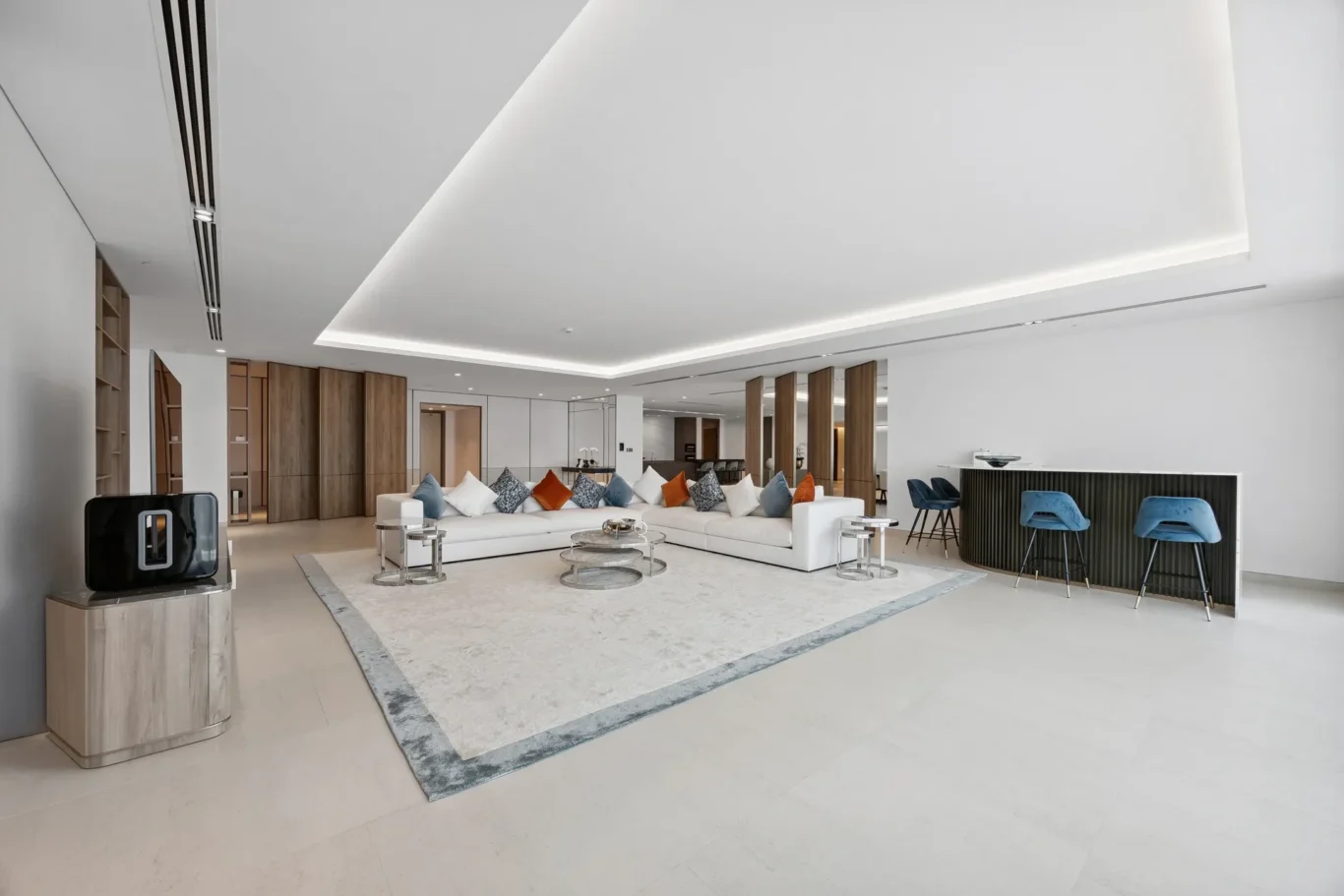 Spacious modern living room with a white sectional sofa, colorful cushions, and glass coffee tables on a large rug backdropped by wood panels and a bar area nearby.