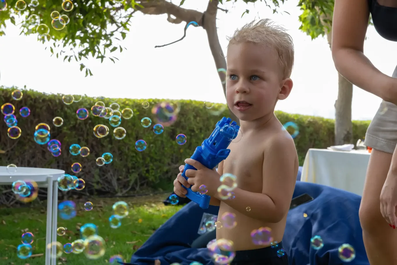Toddler outdoors, shirtless, holding a blue bubble gun while colorful bubbles fill the air near a grassy yard.
