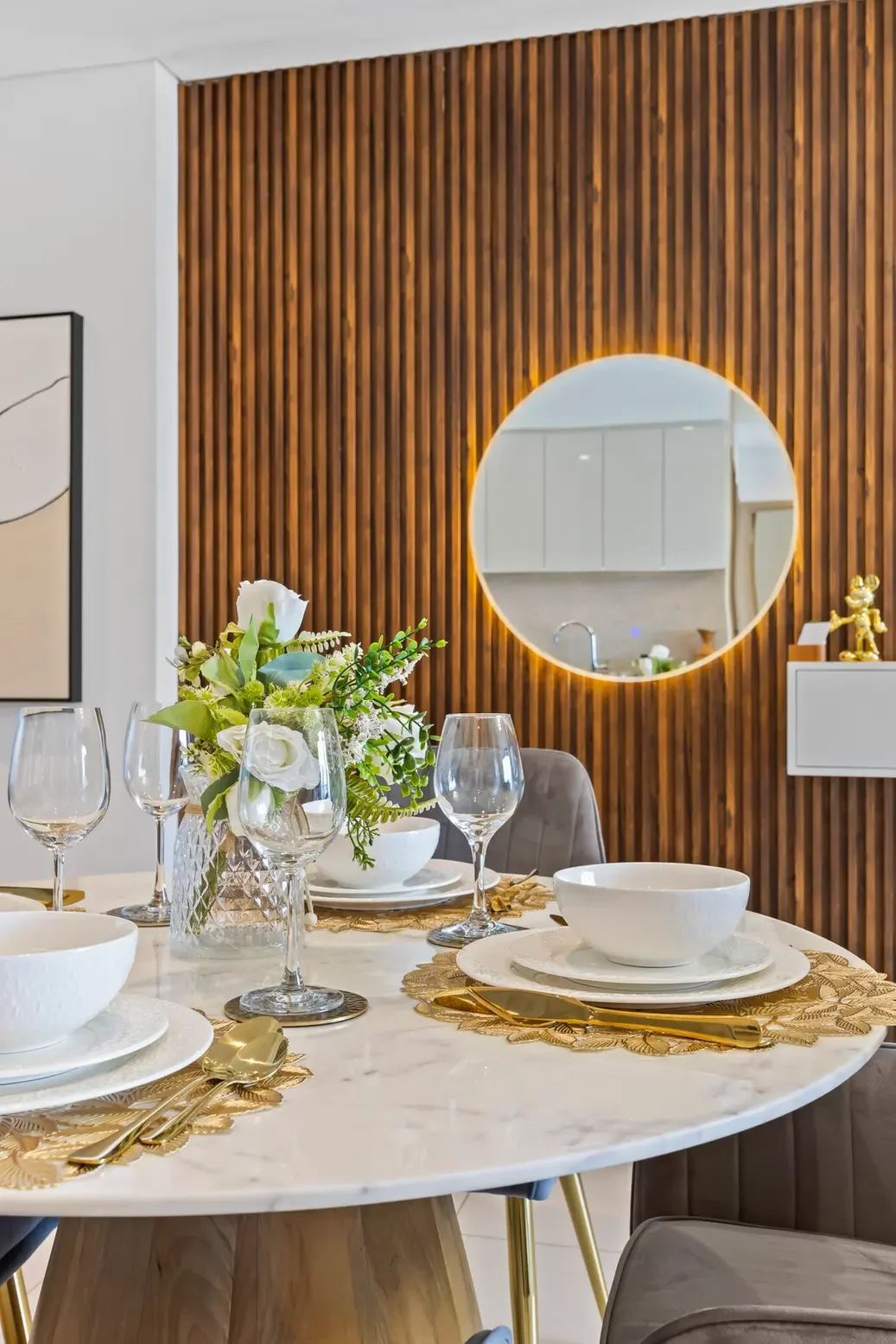 Elegant round marble dining table set for a meal with white dishes, gold flatware, crystal glasses, and a floral centerpiece against a wood-paneled wall with a backlit circular mirror in the background.