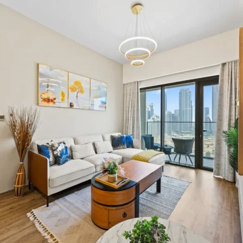 Bright contemporary living room with a white sectional sofa, blue patterned cushions, a round wooden coffee table, and a balcony with city skyline view.