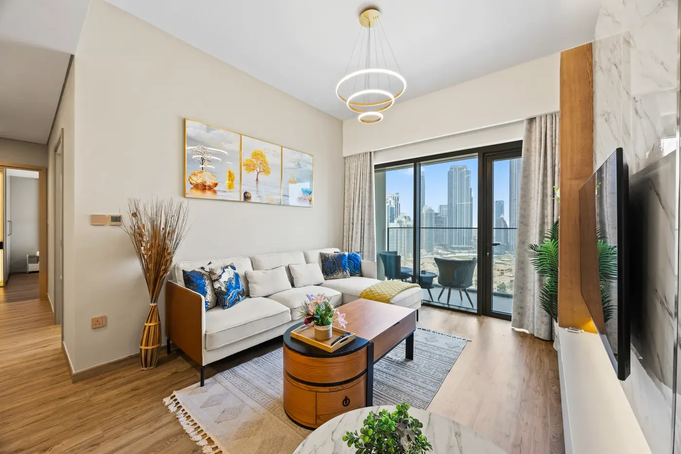 Bright contemporary living room with a white sectional sofa, blue patterned cushions, a round wooden coffee table, and a balcony with city skyline view.