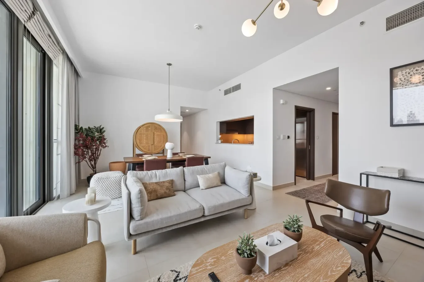 Bright open-concept living room with a gray sofa, brown chairs, and large windows with white curtains; plants and a round coffee table in the foreground.