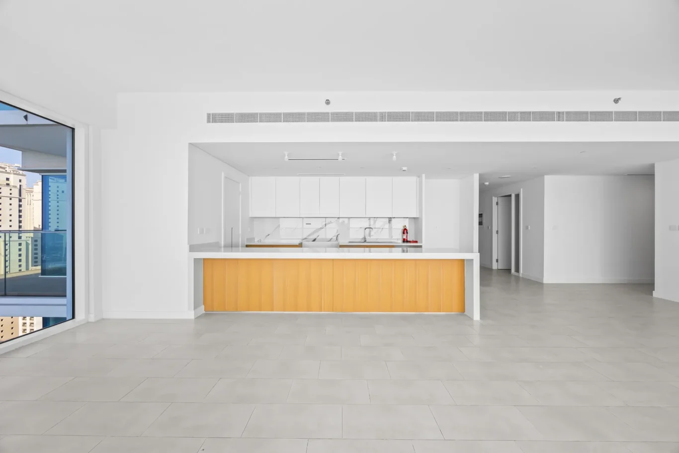 Bright open-plan kitchen with white cabinets, marble backsplash, and a warm yellow island front; large windows reveal a cityscape.