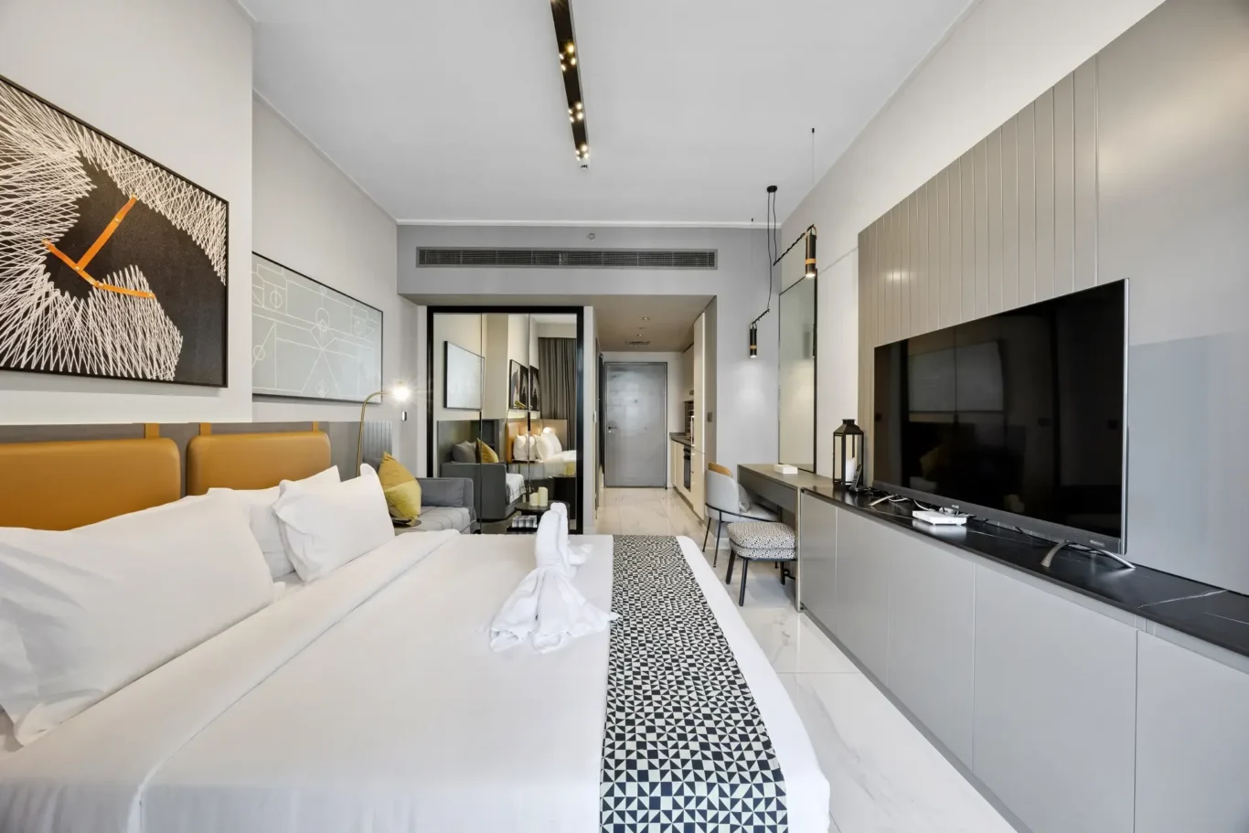 Modern hotel suite with two white beds, orange headboard accents, black-and-white geometric runner, and large flat-screen TV opposite.