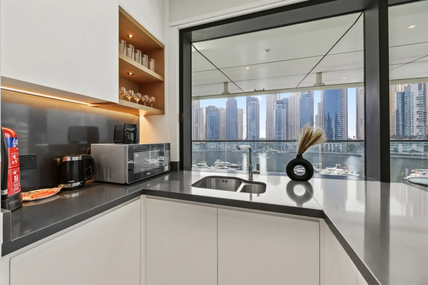 Modern kitchen with white cabinets, dark countertops, kettle and microwave; city skyline visible through a large window.