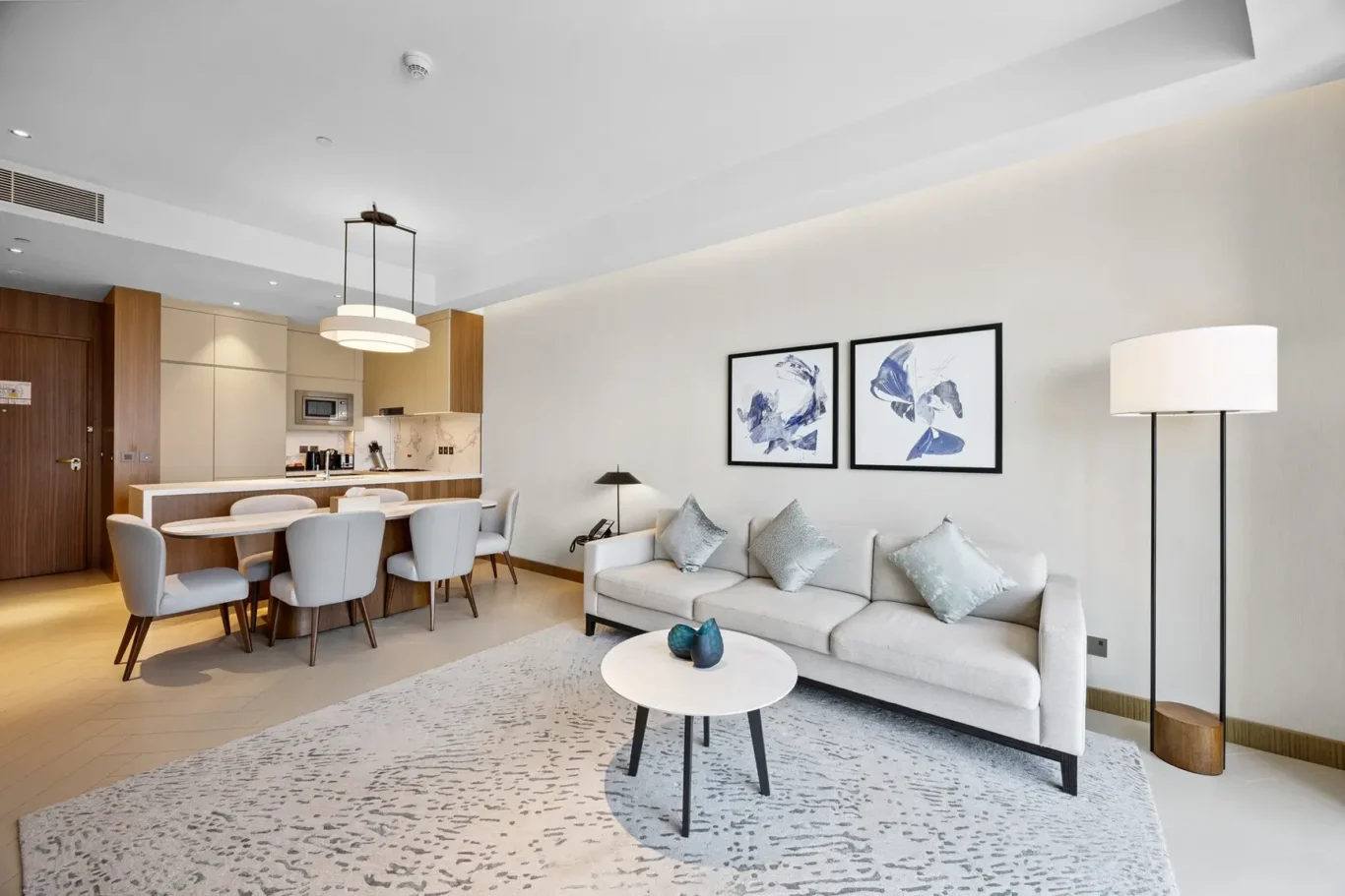 Open-concept living area: cream sofa with pillows, two abstract blue-framed artworks, coffee table, and a kitchen with an island in the background.