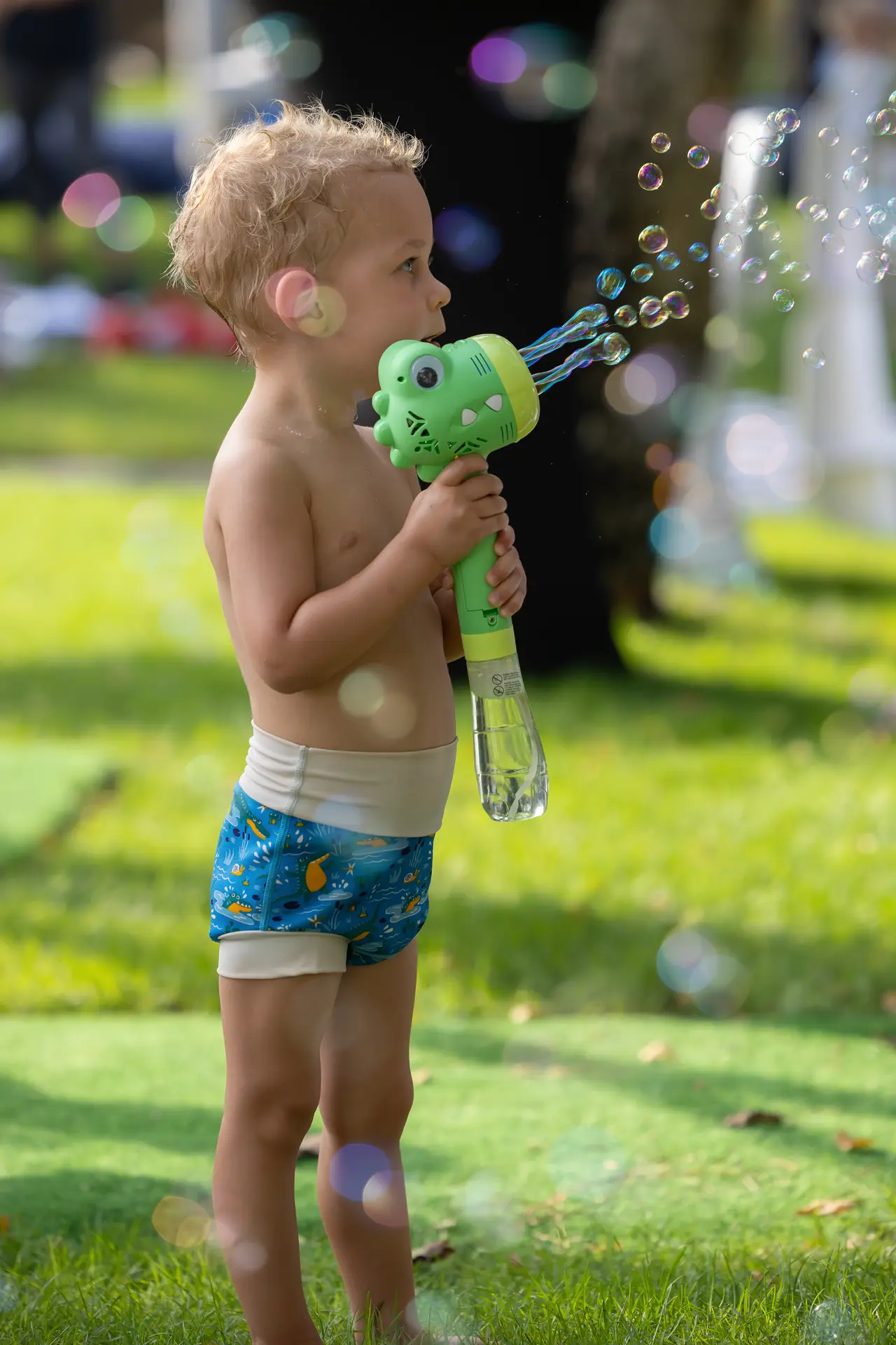 Toddler boy in swim trunks blowing bubbles with a green bubble gun outdoors in a sunny park.