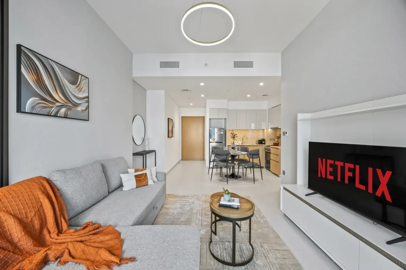 Open-plan living room with a gray sofa, orange throw, round coffee tables, wall art, and a TV displaying Netflix in the foreground; kitchen visible in the background.