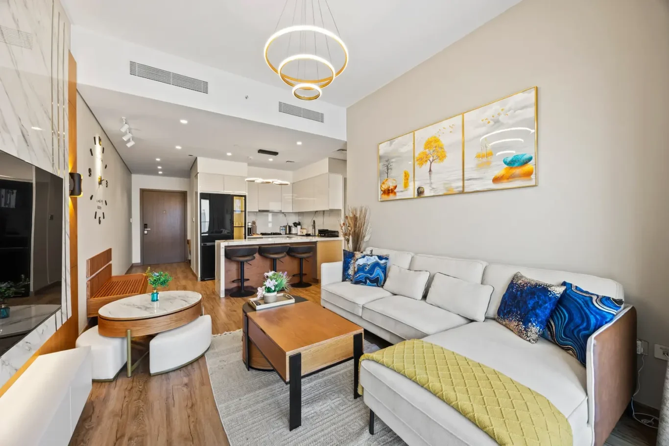 Open-concept living room with a light gray sectional sofa, blue decorative cushions, wooden coffee table, and a kitchen island with three bar stools visible in the background.