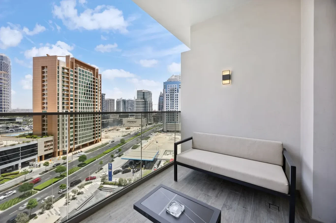 Balcony seating area with a cream cushioned bench and glass railing, overlooking a cityscape of tall buildings and a busy street.