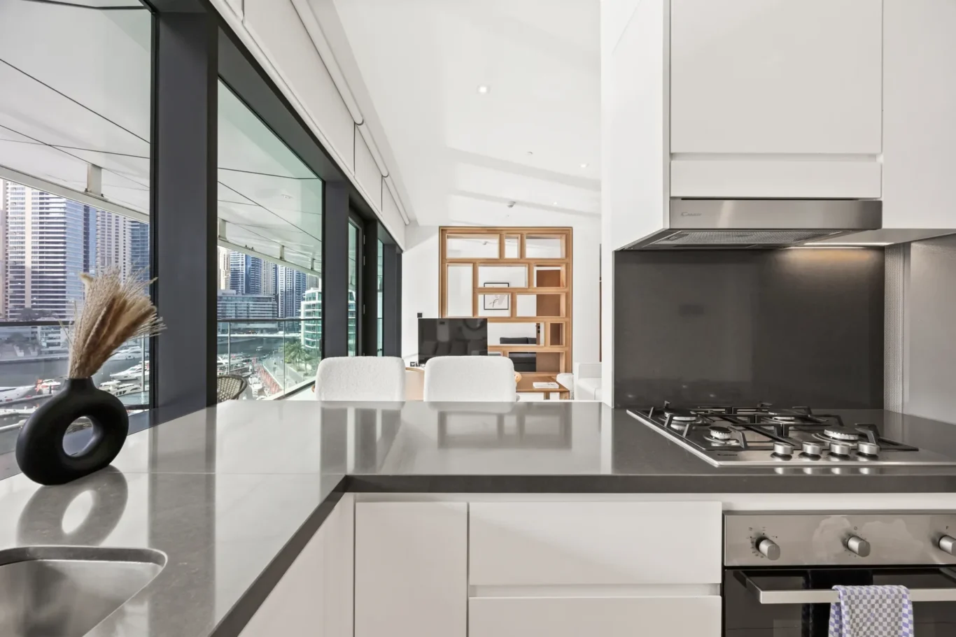 Modern kitchen with white cabinets, a stainless steel countertop, and a gas range, overlooking a city skyline through large windows.