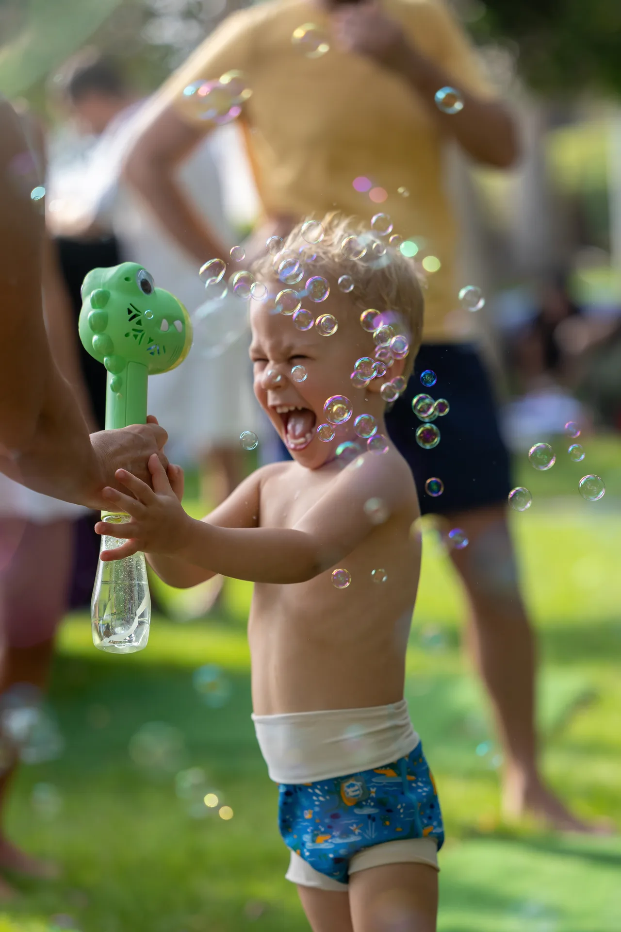 Young child wearing swim trunks blows bubbles with a green bubble wand outdoors, smiling face in motion.