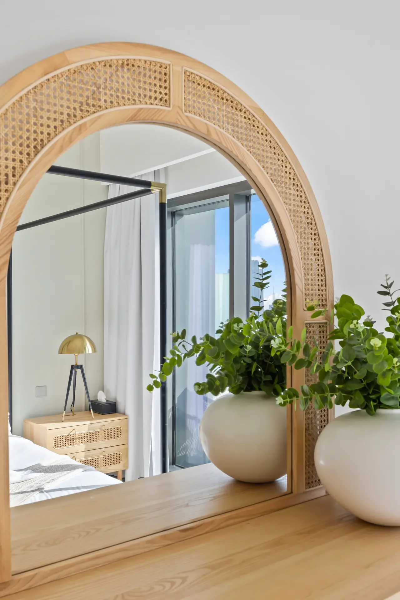 Round wicker-framed mirror above a light wooden dresser, with green plants and a brass lamp by a glass door leading to a balcony.