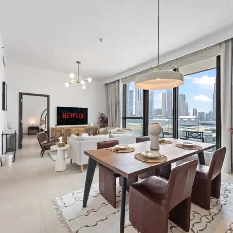 Living and dining area with white sofas, TV showing Netflix, and floor-to-ceiling windows overlooking a city skyline.