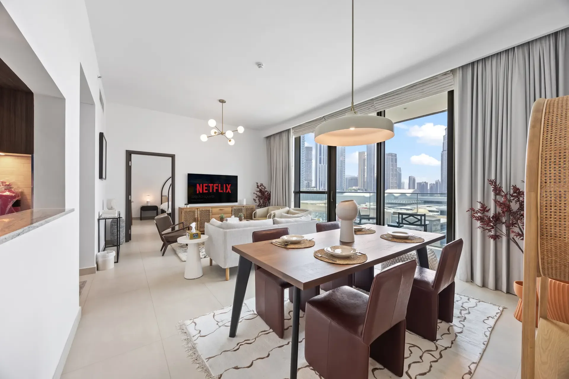 Living and dining area with white sofas, TV showing Netflix, and floor-to-ceiling windows overlooking a city skyline.