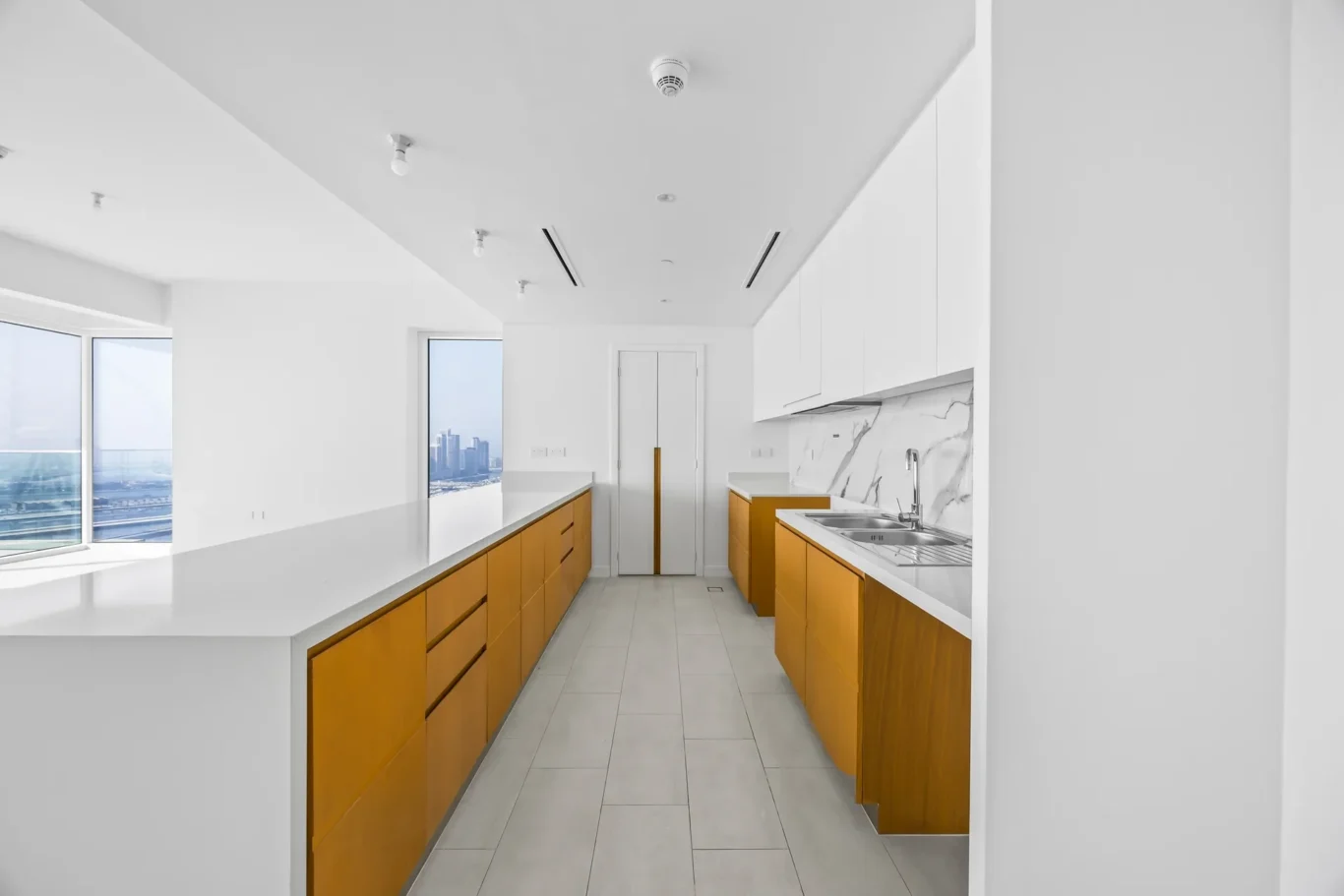 Modern kitchen with white upper cabinets, orange lower cabinets, a long white countertop island, and a city view through large windows on the left.