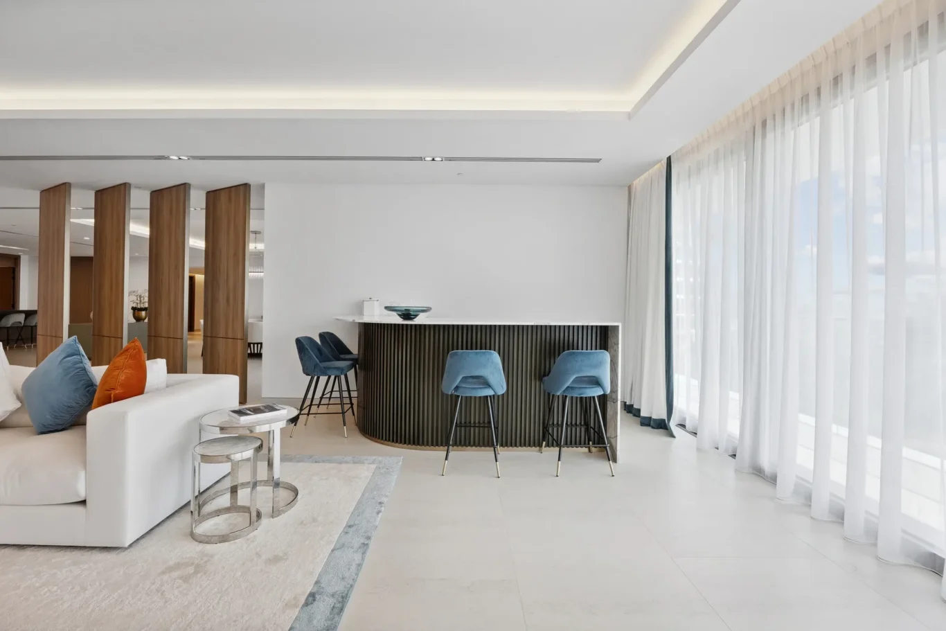Bright hotel lobby with white seating, blue bar stools at a curved dark bar, and sheer curtain windows.