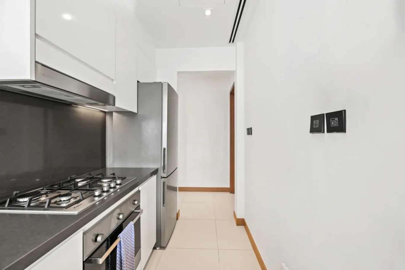 Modern white kitchen with stainless steel appliances and a dark countertop along the left; gas stove and oven visible, refrigerator at back, hallway leading to a wooden door on the right.