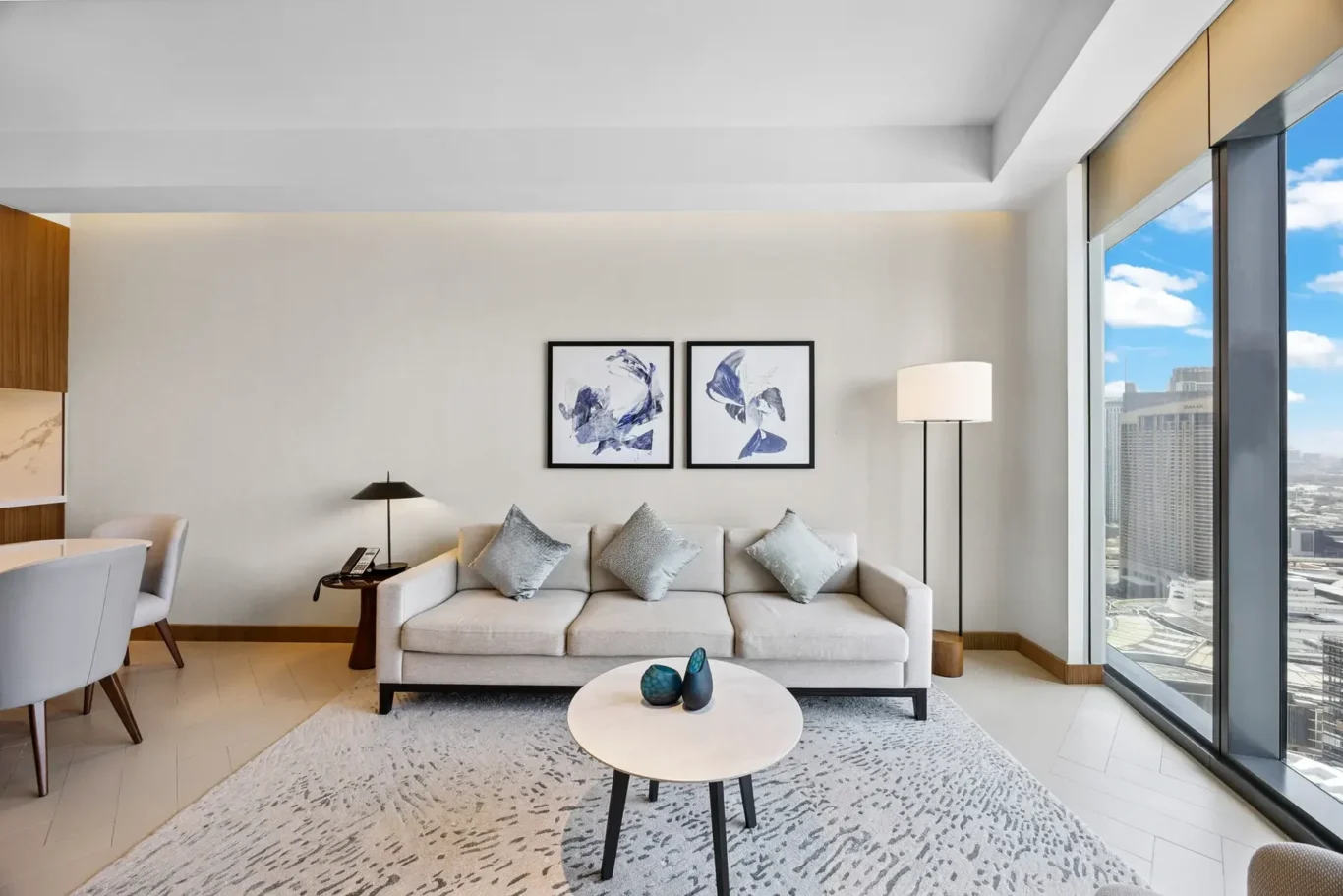 Neutral living room with a beige sofa and five light-blue pillows, two framed abstract prints, and large windows revealing a city skyline outside.