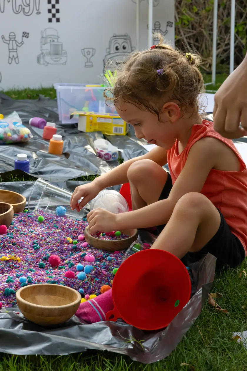 Young girl kneels on a tarp, sorting multicolored beads in a wooden bowl during an outdoor crafts activity.