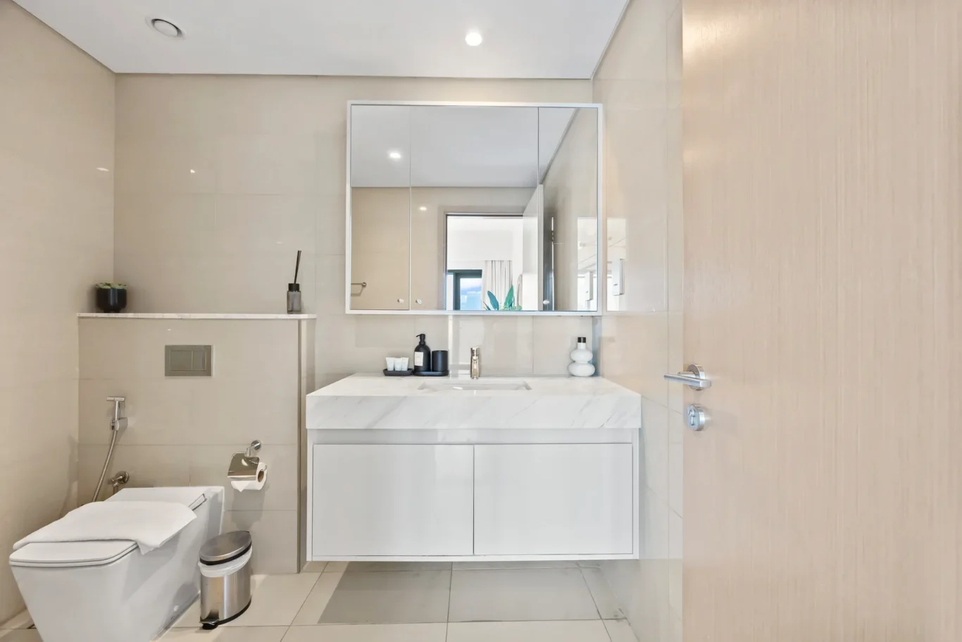 Modern bathroom with a white marble vanity, large mirrored cabinet, beige tiled walls, and chrome fixtures in a bright, minimalist space.
