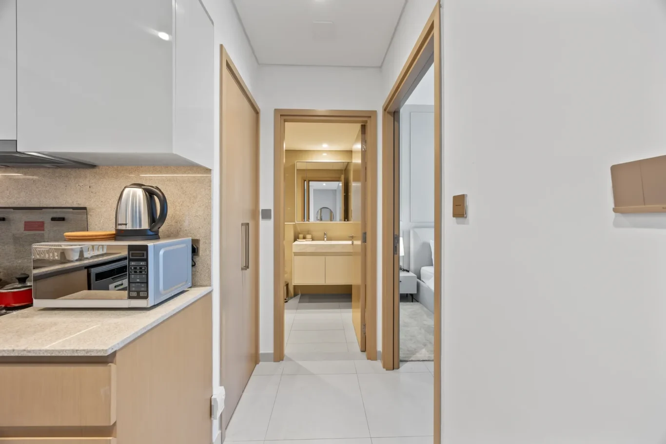 Modern kitchen with white cabinets, a kettle and microwave on a speckled countertop, open to a hallway leading to other rooms.