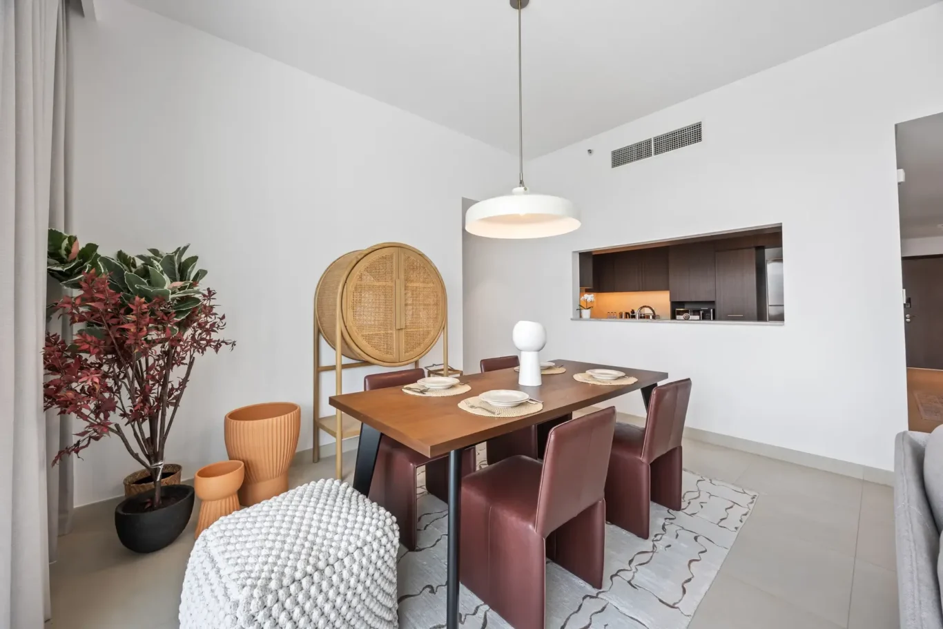 Modern dining area with a wooden table, brown chairs, and a large white pendant light; open kitchen pass-through in the background.
